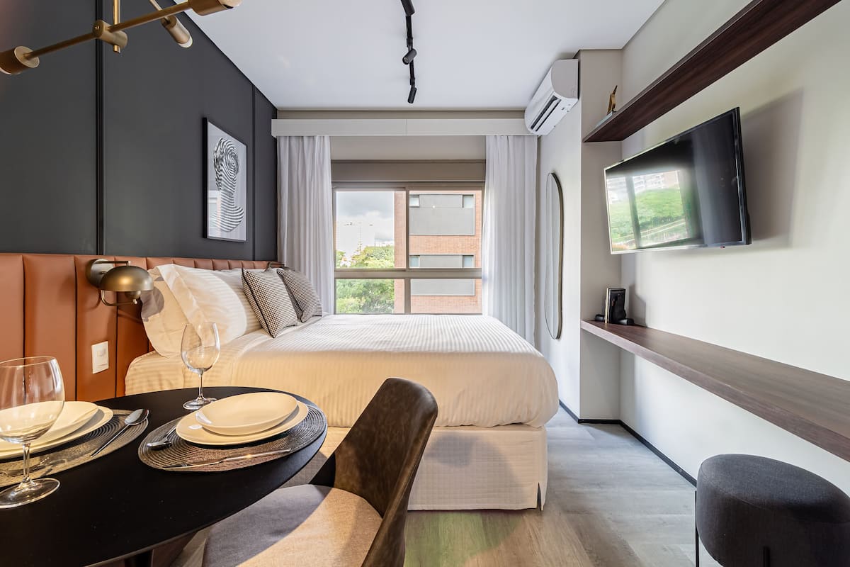 A modern hotel room with a large bed, a dining table set with plates and glasses, a flat-screen TV mounted on the wall, and a window with a view of buildings and greenery.