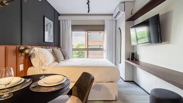 Habitación de hotel moderna con una cama grande, una mesa de comedor con platos y vasos, un televisor de pantalla plana montado en la pared y una ventana con vistas a edificios y vegetación.