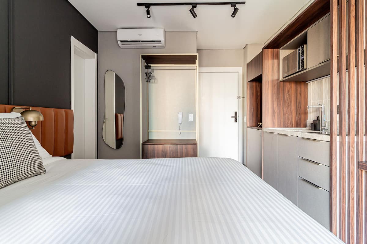A modern hotel room with a large bed, a built-in bench, a mirror, and a kitchenette.