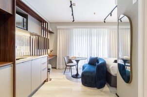 Quarto de hotel moderno com cozinha, mesa de jantar e cadeiras, cama grande com cabeceira azul e um espelho grande na parede.
