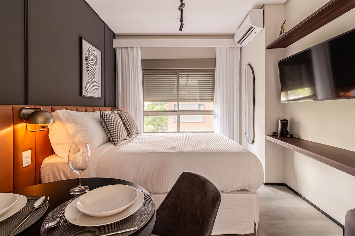 A hotel room with a bed, bedside table with a wine glass, plates, and cutlery. There is a TV mounted on the wall and a window with blinds.