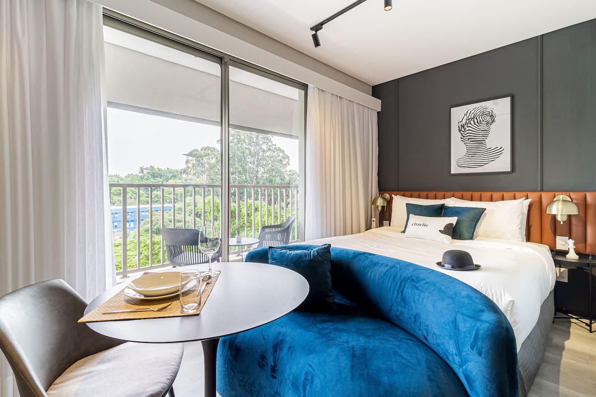 A modern hotel room with a large bed, a blue sofa, a dining table with a placemat, and a balcony with a view of greenery.