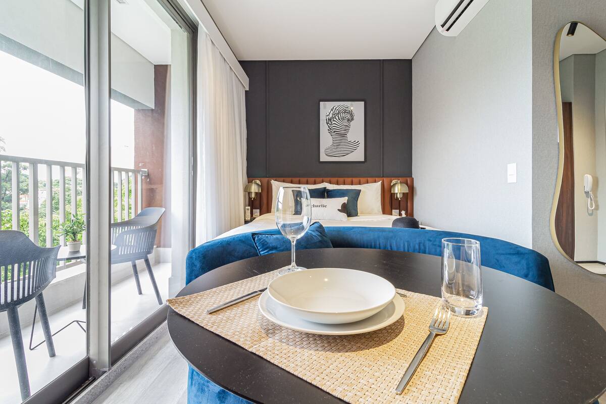 A hotel room with a bed, a dining table with a plate, a glass, and a fork. There is a balcony with chairs and a potted plant.