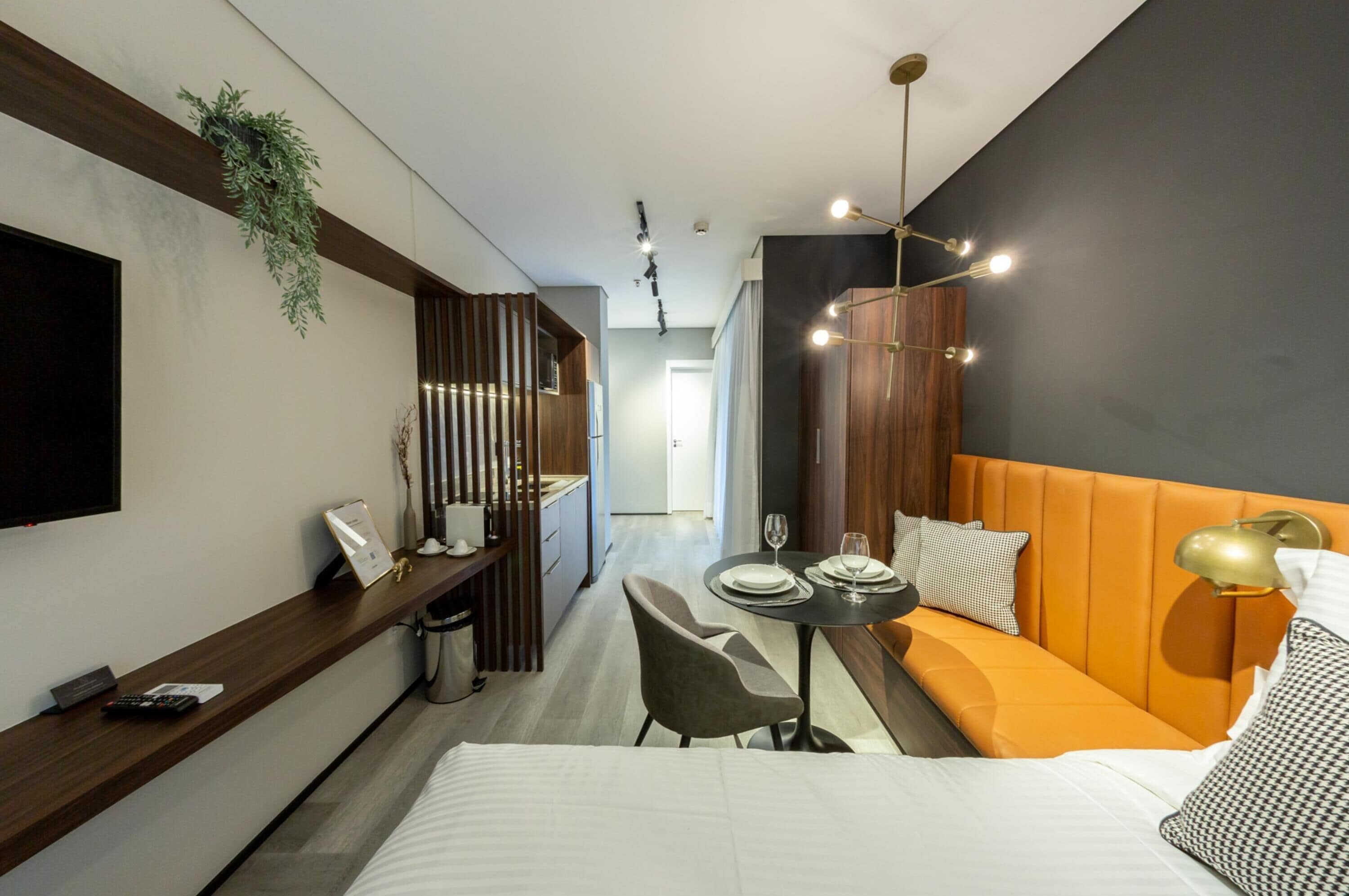 A modern hotel room with a bed, a dining table with chairs, a sofa, a TV mounted on the wall, and a plant.