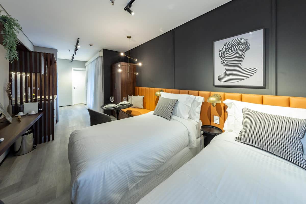 A modern hotel room with two beds, a desk, a chair, a small table with a lamp, and a large abstract painting on the wall.