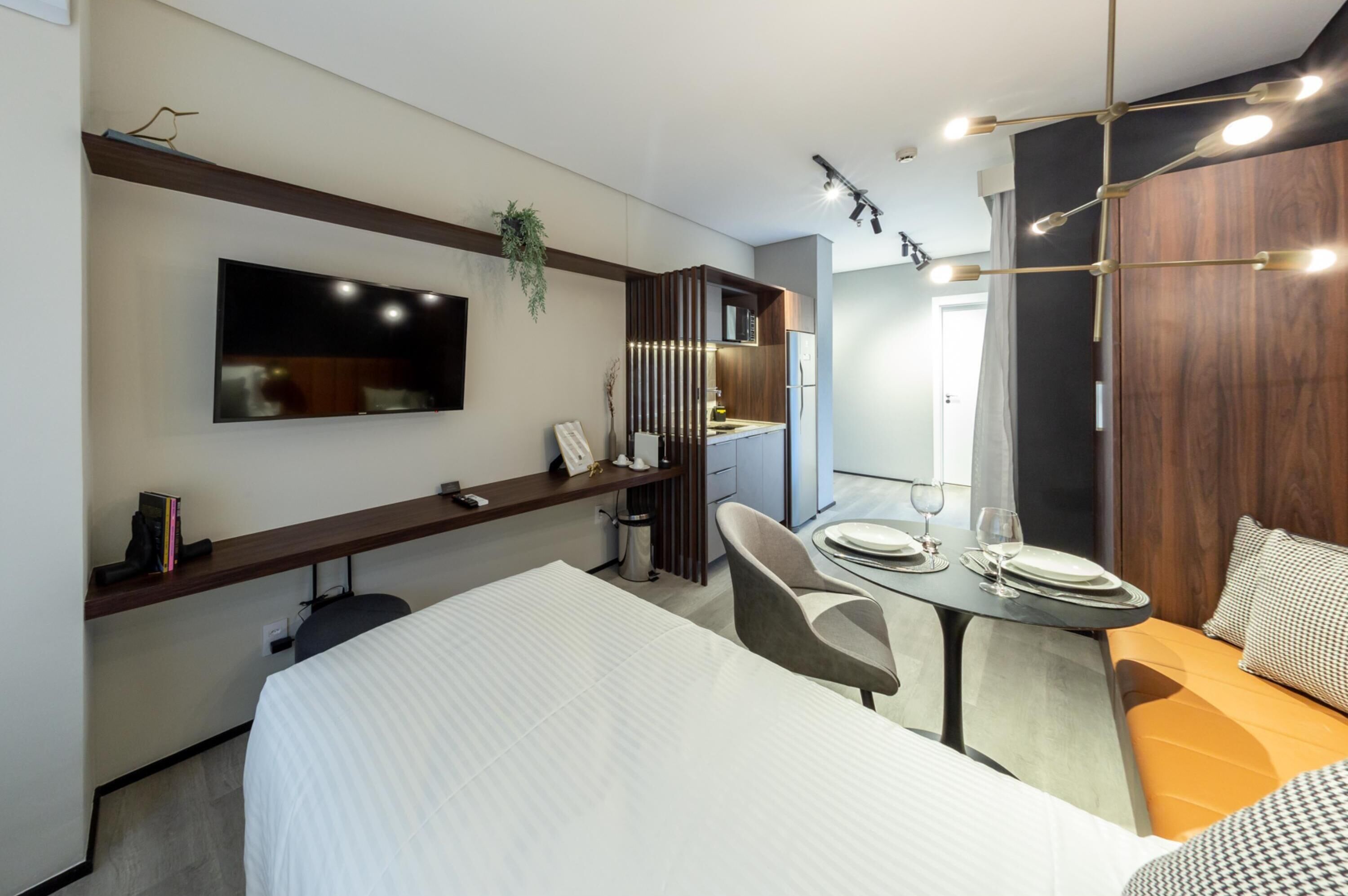 Una habitación de hotel moderna con una cama, una mesa de comedor con sillas, un televisor montado en la pared y una pequeña cocina.