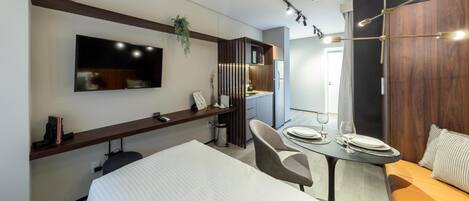 A modern hotel room with a bed, a dining table with chairs, a TV mounted on the wall, and a small kitchen area.