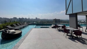 A rooftop pool area with lounge chairs and a table, overlooking a city skyline.