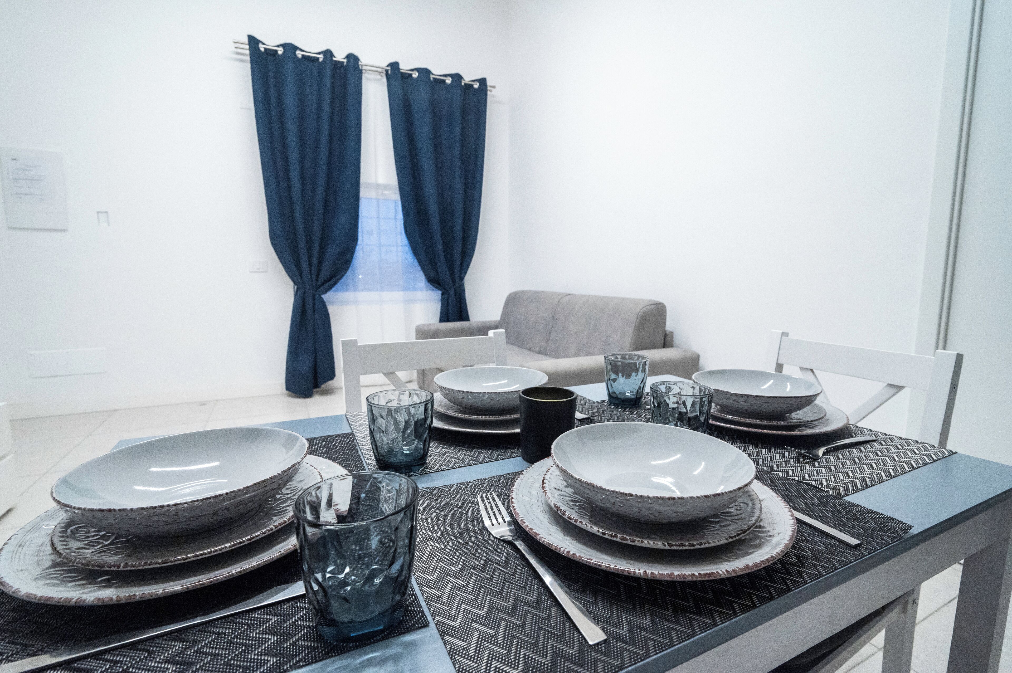 Uma área de jantar com uma mesa posta para quatro pessoas, incluindo pratos, tigelas, copos e talheres.