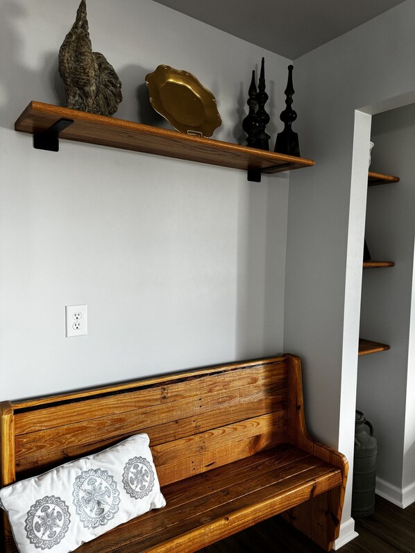 A wooden bench with a decorative pillow, a wall-mounted shelf with a decorative plate and a sculpture, and a white wall with a power outlet. - Cozy Southern Charm in Houma (Houma)