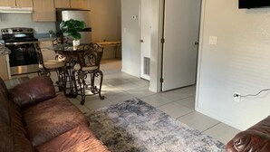 Ein Wohnzimmer mit braunem Ledersofa, ein Essbereich mit Tisch und Stühlen, eine Küche mit Schränken und Geräten sowie eine Tür zu einem anderen Raum.