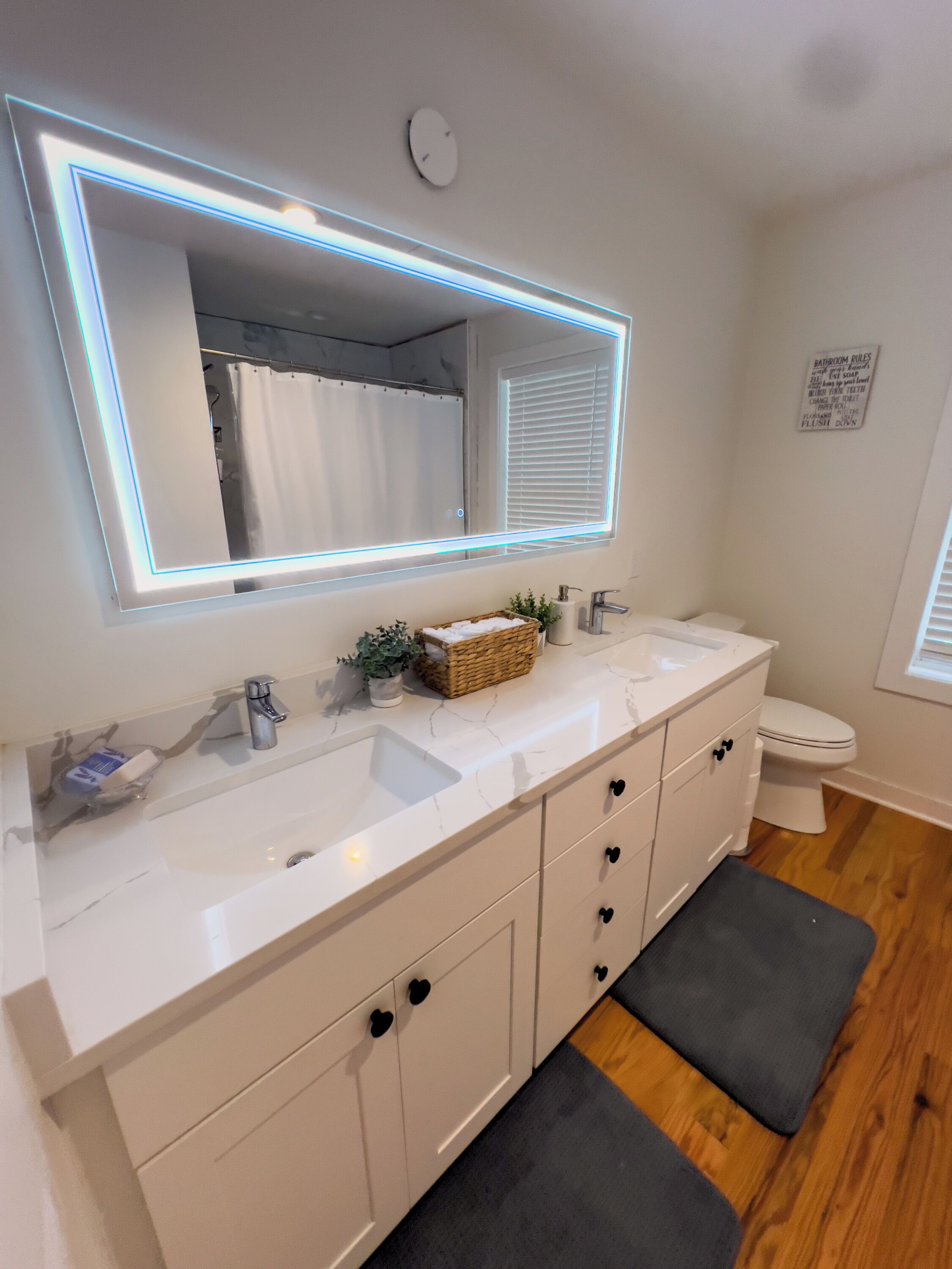 Un baño con tocador blanco, un espejo grande con iluminación LED, inodoro blanco y piso de madera.