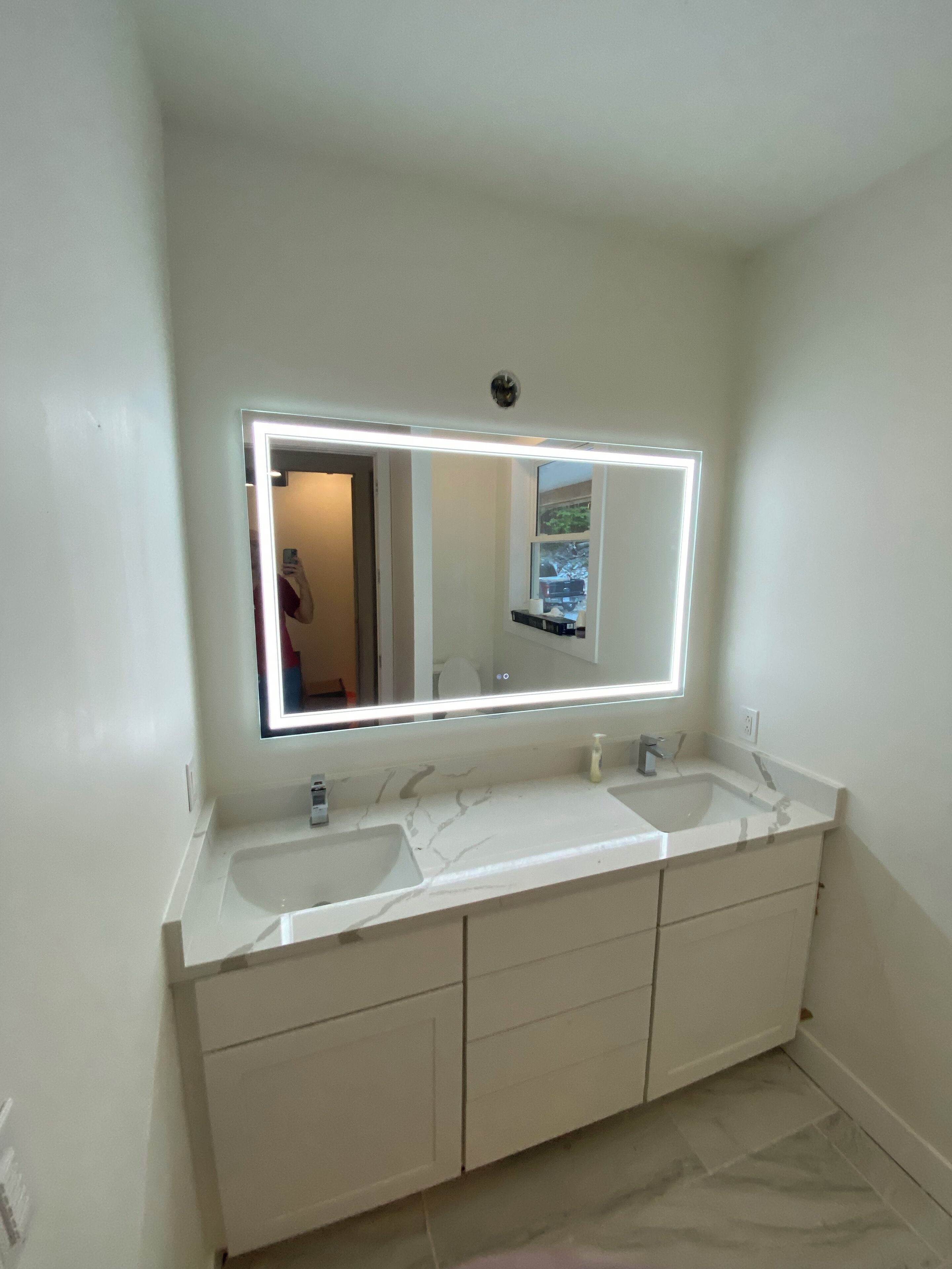 A bathroom with a double sink vanity, a large mirror with integrated lighting, and a plain tiled floor.