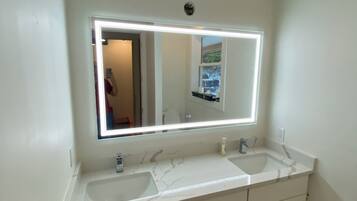 A bathroom with a double sink vanity, a large mirror with integrated lighting, and a plain tiled floor.