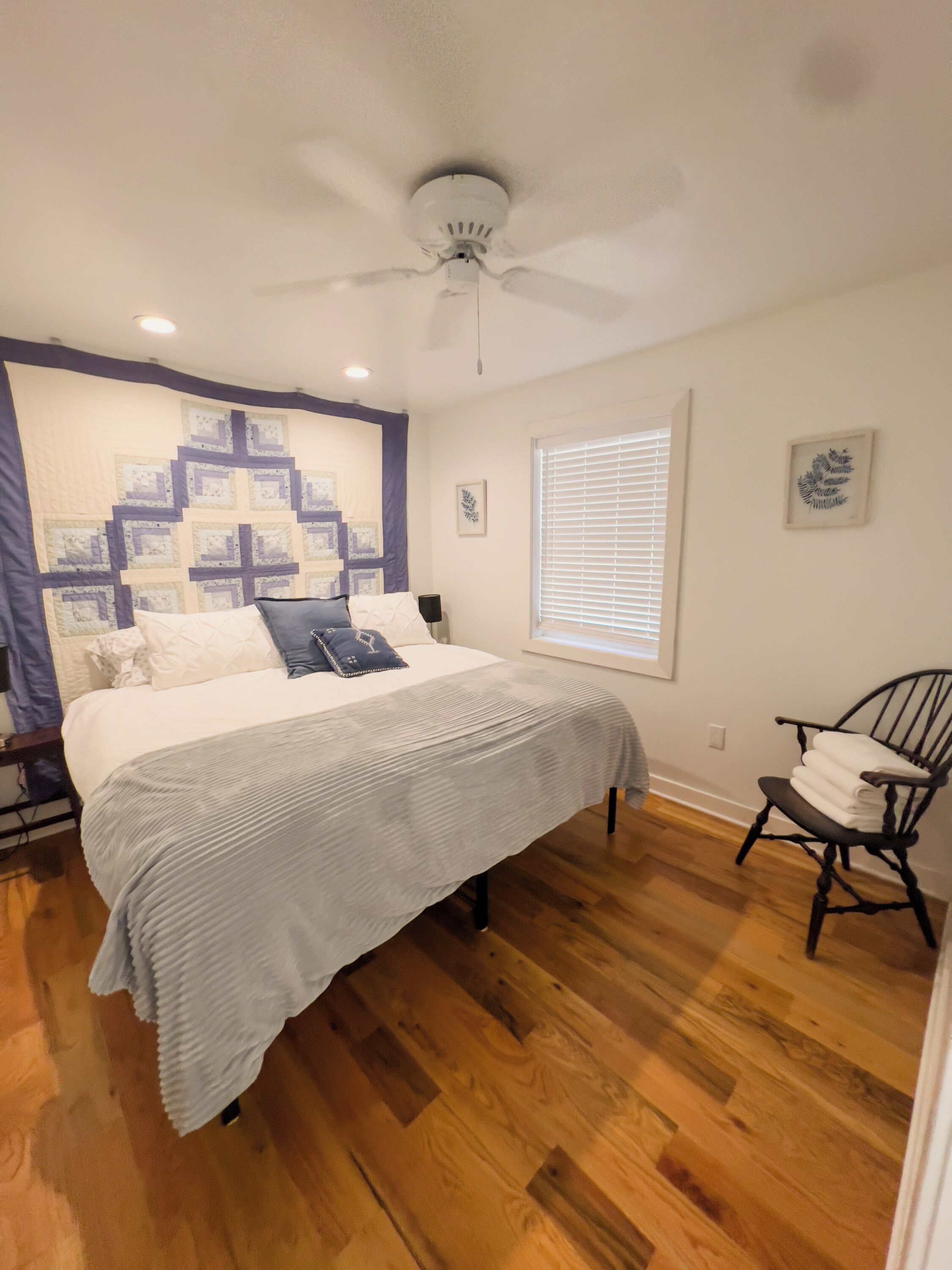 A bedroom with a bed, a ceiling fan, a window with blinds, a chair, and a quilt on the wall.