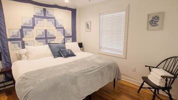 A bedroom with a bed, a ceiling fan, a window with blinds, a chair, and a quilt on the wall.