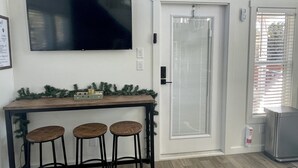 A room with a flat-screen TV mounted on the wall, a wooden console table with three stools, a door with a frosted glass panel, and a small refrigerator.
