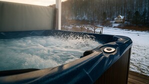 A hot tub on a wooden deck with a snowy landscape and a house in the background.
