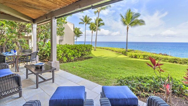 Uma área externa coberta com vista para o oceano, com uma mesa, cadeiras e um pequeno jardim com palmeiras.