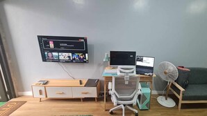 A room with a desk, two monitors, a chair, a sofa, and a wall-mounted TV.