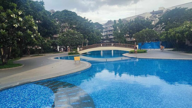 A swimming pool with a tiled edge, surrounded by trees and a pedestrian bridge.
