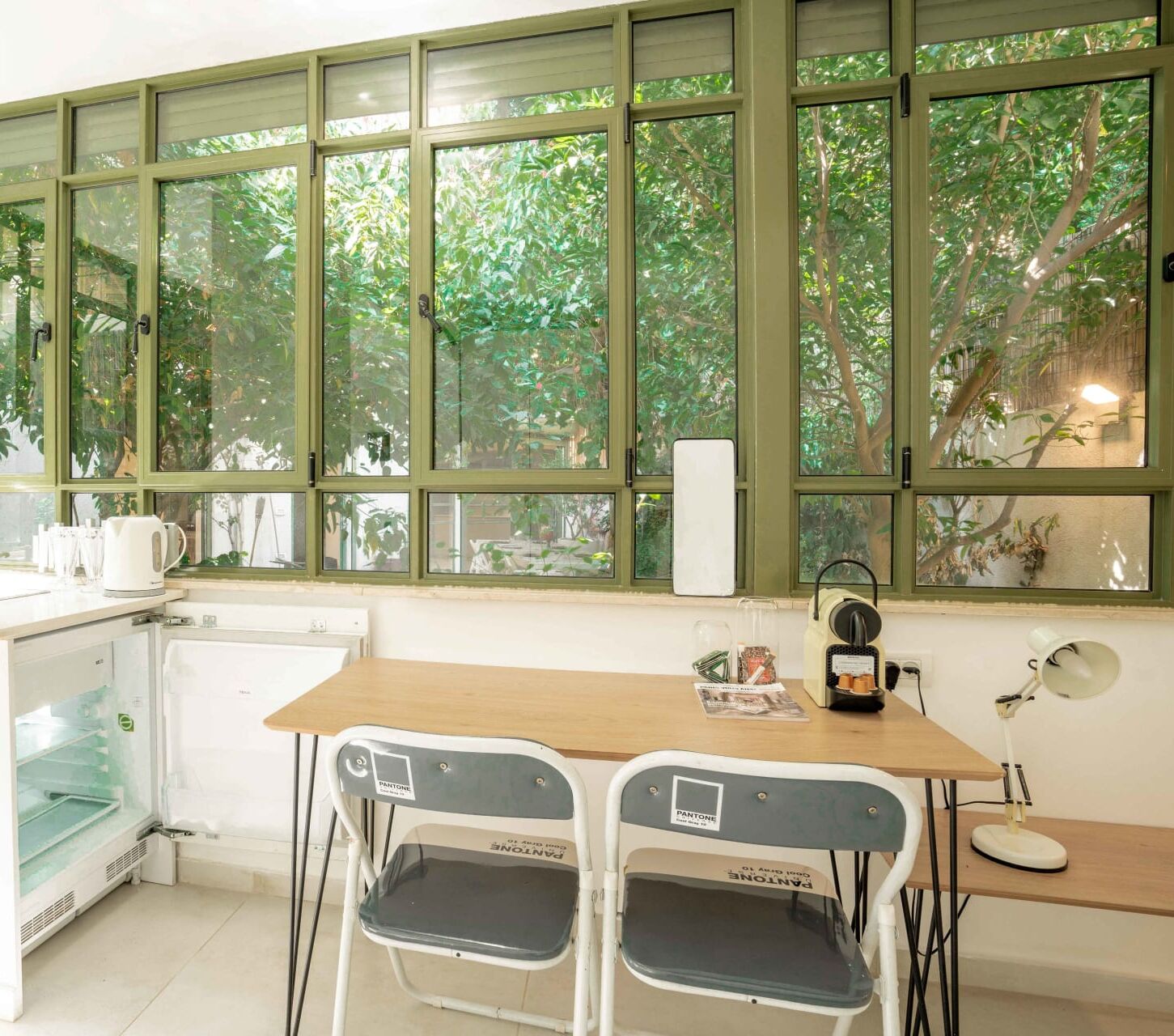 A compact kitchen with a window overlooking greenery, a small table, two chairs, and a refrigerator.