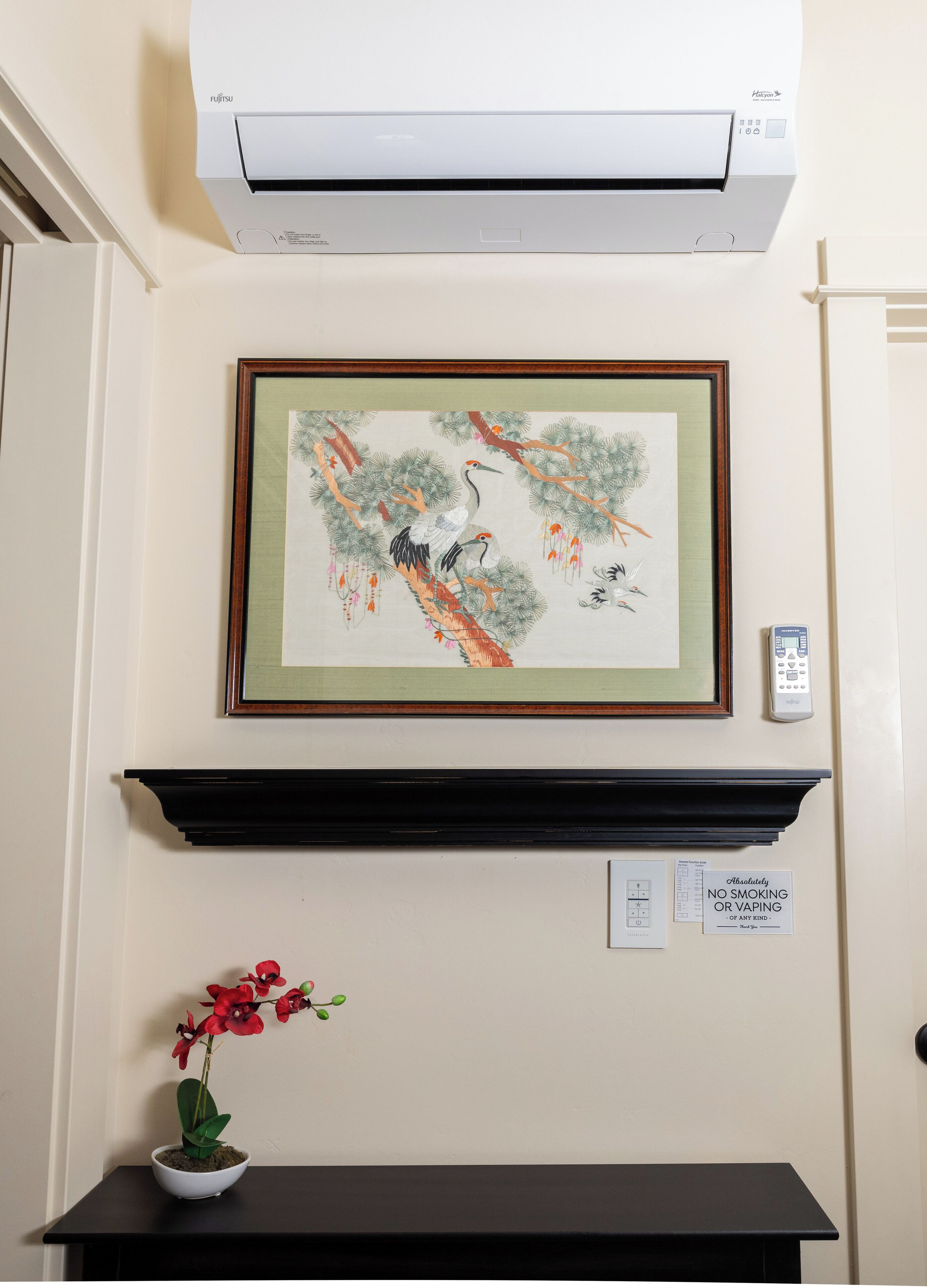 Un climatiseur fixé au mur, un tableau encadré, une télécommande, un panneau indiquant qu’il est interdit de fumer et un pot d’orchidée.