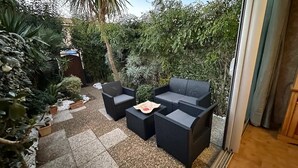 Eine Terrassengruppe im Freien mit Korbmöbeln, darunter eine Couch und zwei Sessel, umgeben von verschiedenen Blumentöpfen und einem kleinen Tisch.