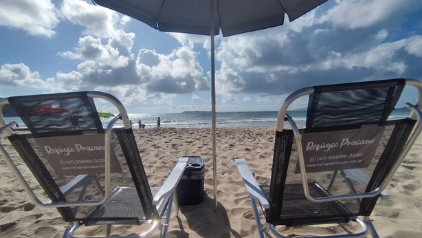 Two beach chairs with a beach umbrella and a cooler on a sandy beach. - Refúgio Praiano (Bombinhas)