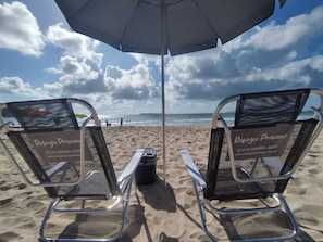Zwei Liegestühle mit Sonnenschirm und Kühlbox am sandigen Strand.