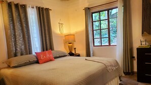 A bedroom with a large bed, a bedside lamp, a window with a view of greenery, and a macrame wall hanging.