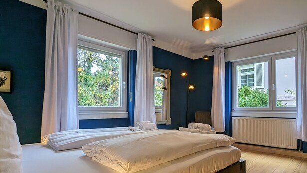 Un dormitorio con una cama grande, dos ventanas con cortinas, un radiador y una lámpara colgante.