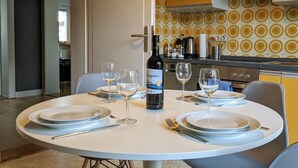 Una mesa de comedor dispuesta con platos, copas de vino y una botella, con una cocina al fondo.