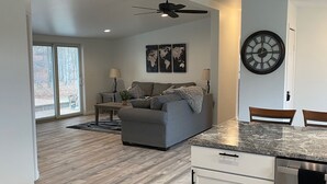 A spacious living area with a grey sofa, a dining table, and a kitchen with white cabinets and a stainless steel dishwasher. - Cozy Country home just 5 miles to Boyne City/8 miles to Boyne Mountain (Boyne City)