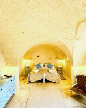 Quarto com arco de pedra, cama de casal com almofadas, mesas de cabeceira, cômoda e uma cadeira.