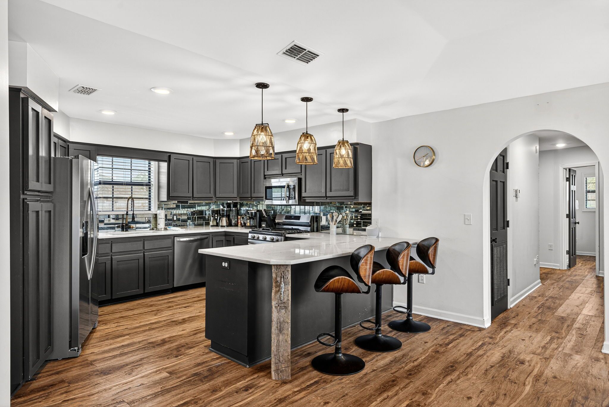 A modern kitchen with dark cabinets, a central island, bar stools, pendant lighting, and a view to the living area.