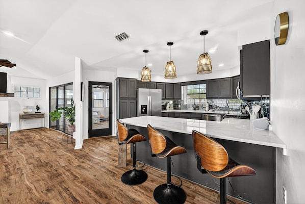 A modern kitchen with a central island, bar stools, pendant lighting, and a view into the dining area.