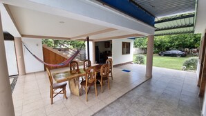 A covered patio area with a wooden table and chairs, a hammock, and a view of a garden and parked car.