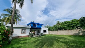 A modern two-story house with a blue roof and white walls, surrounded by a well-maintained lawn and palm trees.