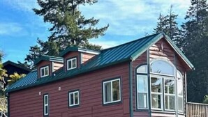 A red wooden cabin with a green roof, a large window, and a deck. - The Lighthouse Cabin #60 (Gold Beach)