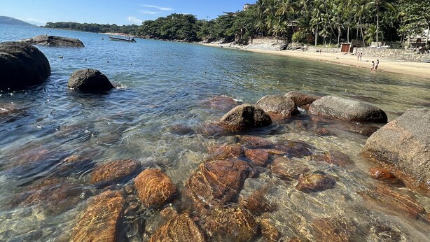 Una playa con agua cristalina, rocas y una colina boscosa al fondo.