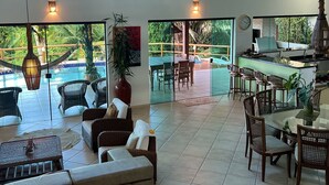 Un espacio interior amplio con techo alto, zona de comedor con mesas y sillas redondas, sala de estar con sofás y una mesa de centro, y vistas a una piscina y zonas verdes exteriores.