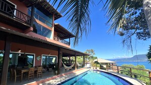 Un área junto a la piscina con vistas al mar, un comedor con mobiliario exterior y un edificio con amplias ventanas y un balcón.