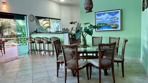 Un comedor con una mesa de vidrio rodeada de sillas de mimbre, una cocina al fondo y un ventanal con vistas a la vegetación.