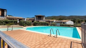 A swimming pool with a wooden deck, surrounded by buildings with a Mediterranean architectural style, set against a backdrop of hills. - Holiday Home 'Manessy' with Sea View, Shared Pool and Air Conditioning (Badesi)