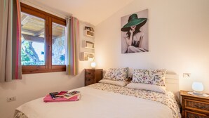 A bedroom with a bed, a nightstand, a lamp, a window with curtains, a wall-mounted shelf with books, and a framed picture. - Holiday Home 'Manessy' with Sea View, Shared Pool and Air Conditioning (Badesi)