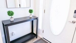 Een moderne inkomhal met een donkere console tafel, twee kleine potplanten, een grote spiegel en een witte deur met een rond raam.