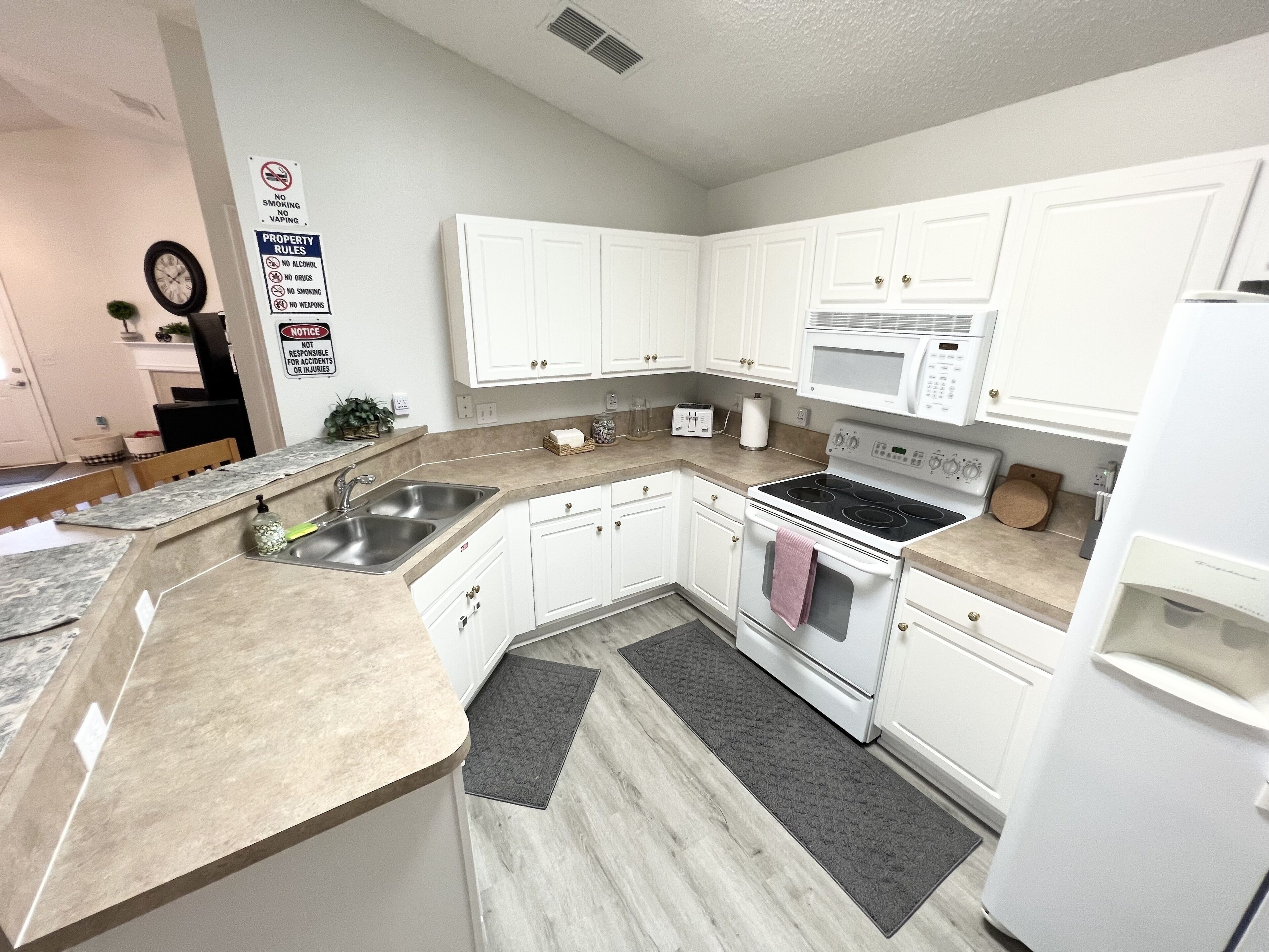 A modern kitchen with white cabinets, a stainless steel sink, a microwave, and a stove. There are two rugs on the floor and a clock on the wall.
