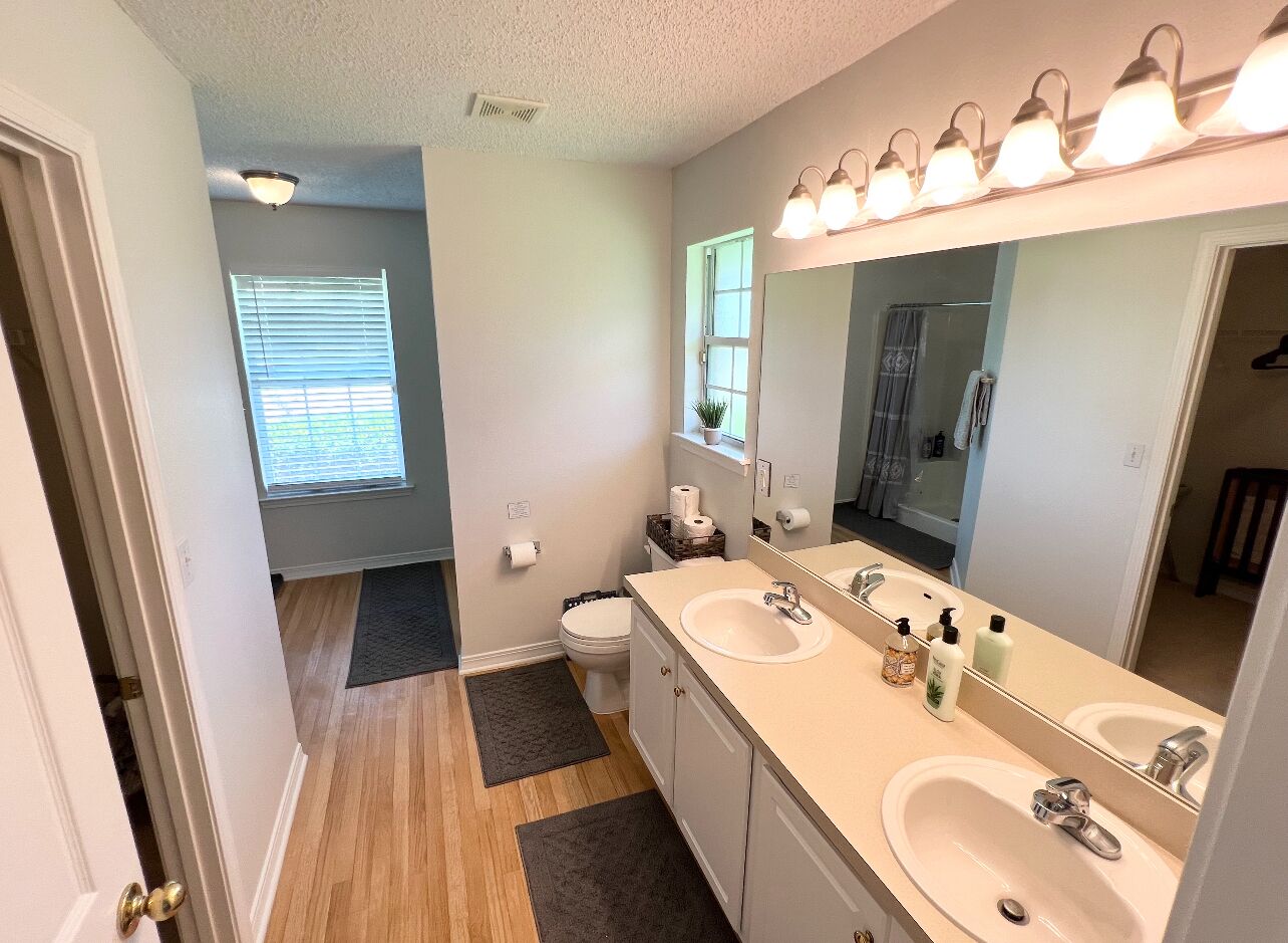 A bathroom with two sinks, a large mirror, and a toilet. There is a window with blinds and a door leading to another room.