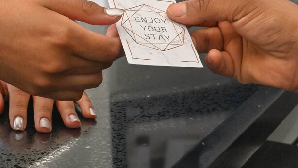 Una mano sosteniendo una tarjeta con la frase 'ENJOY YOUR STAY' y una toalla con el logotipo 'STAY HOME HOTEL'.